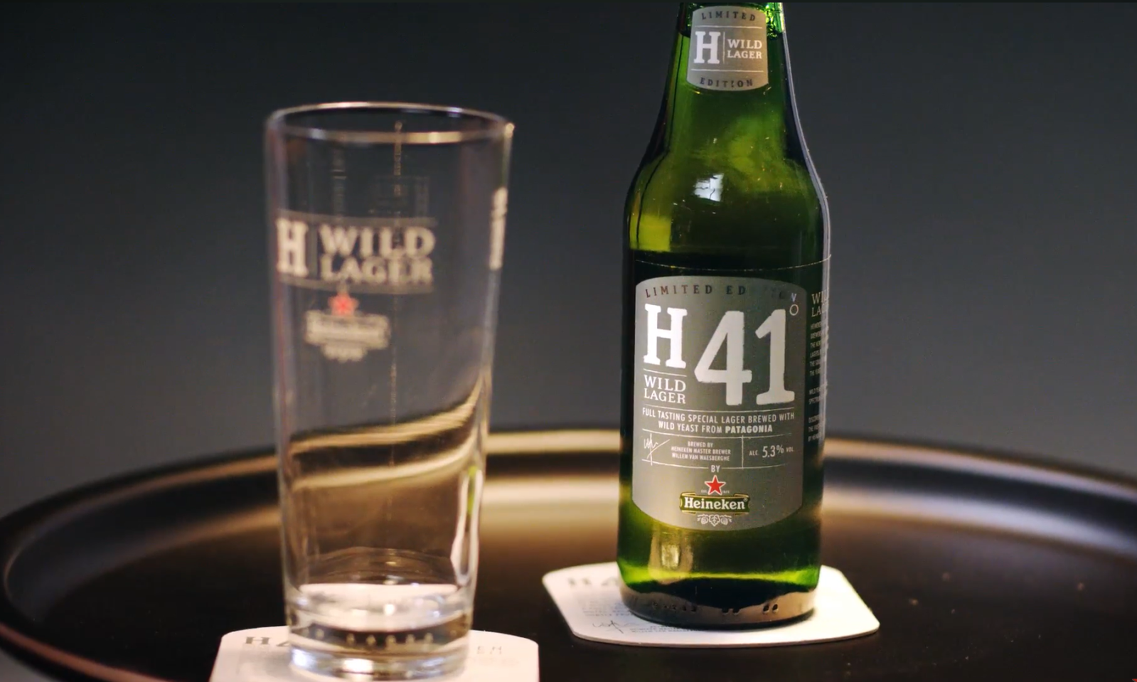 Een fles Heineken Wild Lager H41 naast een leeg glas op een donkere achtergrond.
