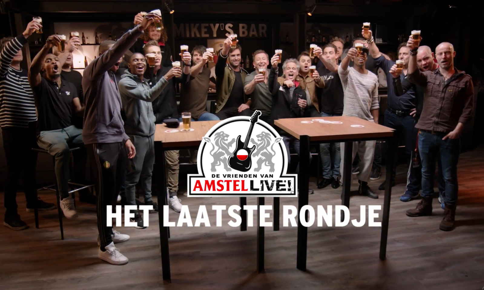 Groep mensen heft gezamenlijk glazen bier in café met logo van Amstel op voorgrond.