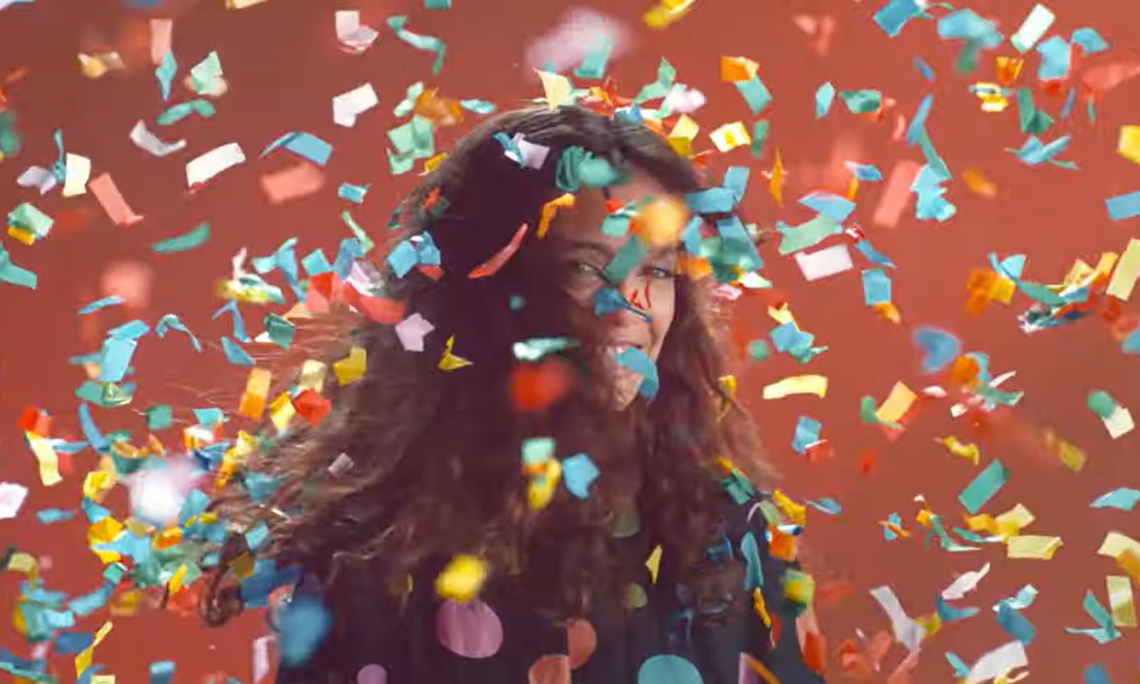 Vrouw glimlachend bedekt met kleurrijke confetti op een rode achtergrond.