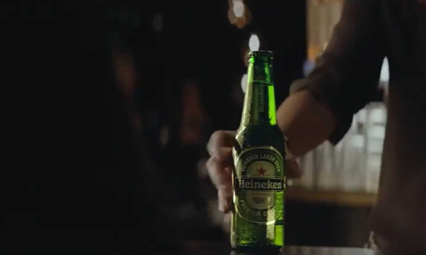 Een hand die een groene fles Heineken vast heeft op een donkere achtergrond.