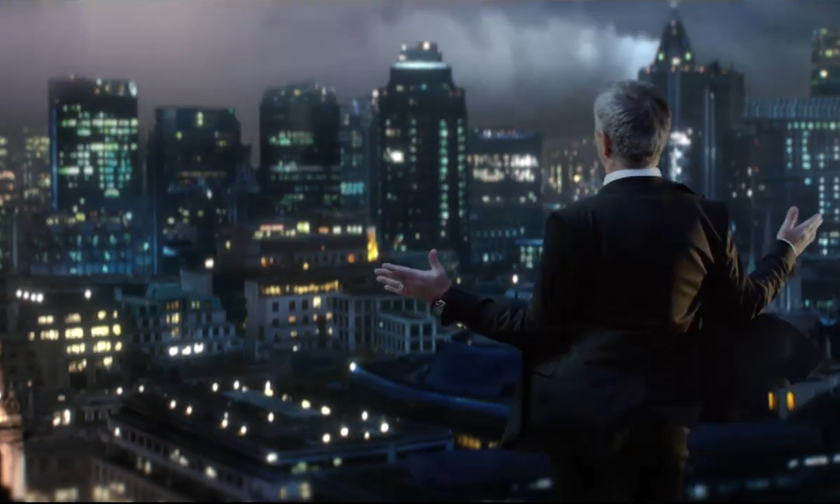 Man in pak met open armen kijkt uit over een verlichte stad bij nacht.