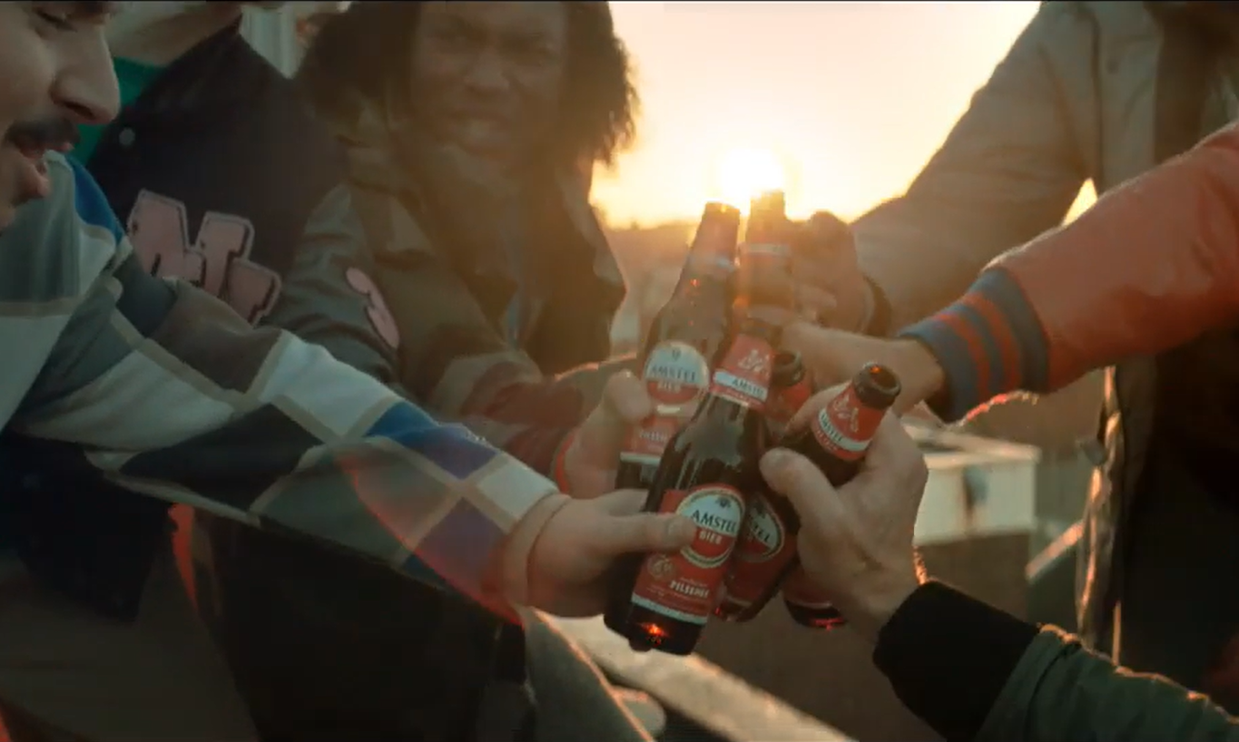 Vier mensen proosten met flesjes bier bij zonsondergang op een stadsdakterras.