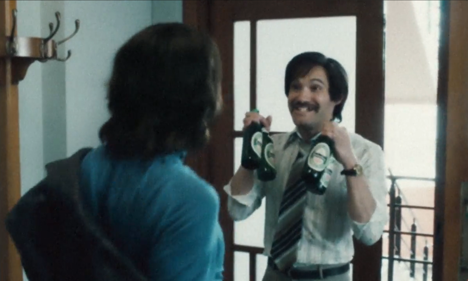 Twee mannen in gesprek bij een deur, een man houdt twee flessen bier omhoog.