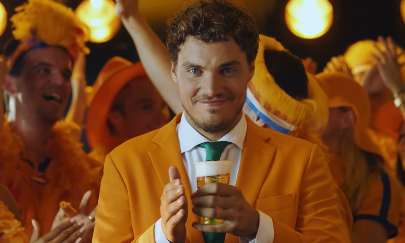 Man in oranje pak klapt met een biertje in zijn hand te midden van een juichende menigte.