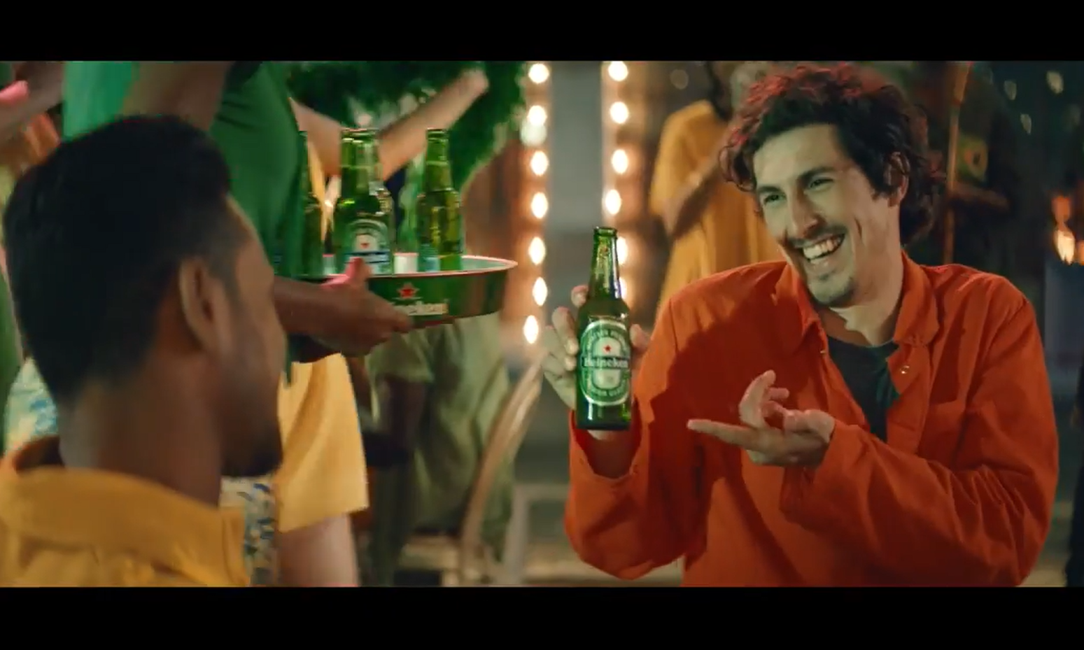 Lachende man in oranje shirt wijst naar fles Heineken tijdens feest.
