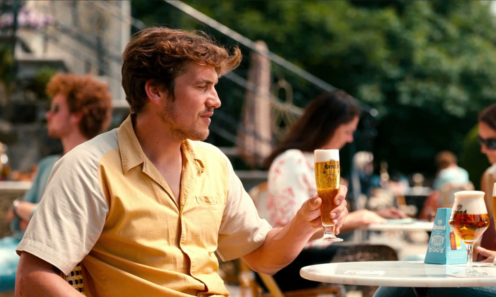 Man houdt een glas bier vast op een zonnig terras met mensen op de achtergrond.