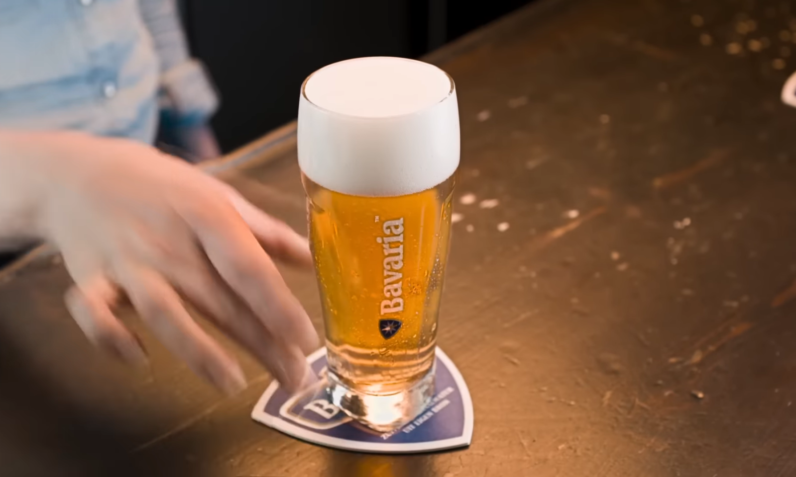 Hand reikt naar een glas bier op een houten tafel met een bierviltje.