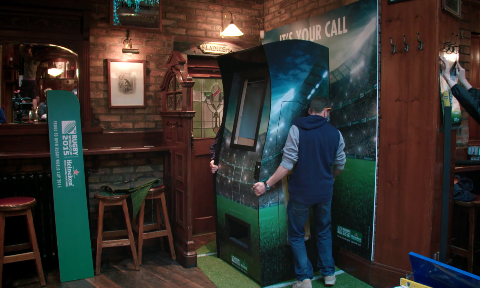 Twee mannen verplaatsen een grote rugbythema-machine in een pub.