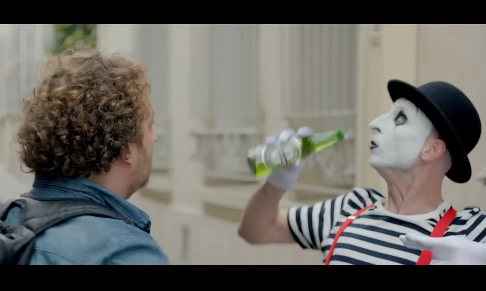Een man in een mime outfit met wit gezicht probeert te drinken uit een groene fles voor een andere man met een rugzak.