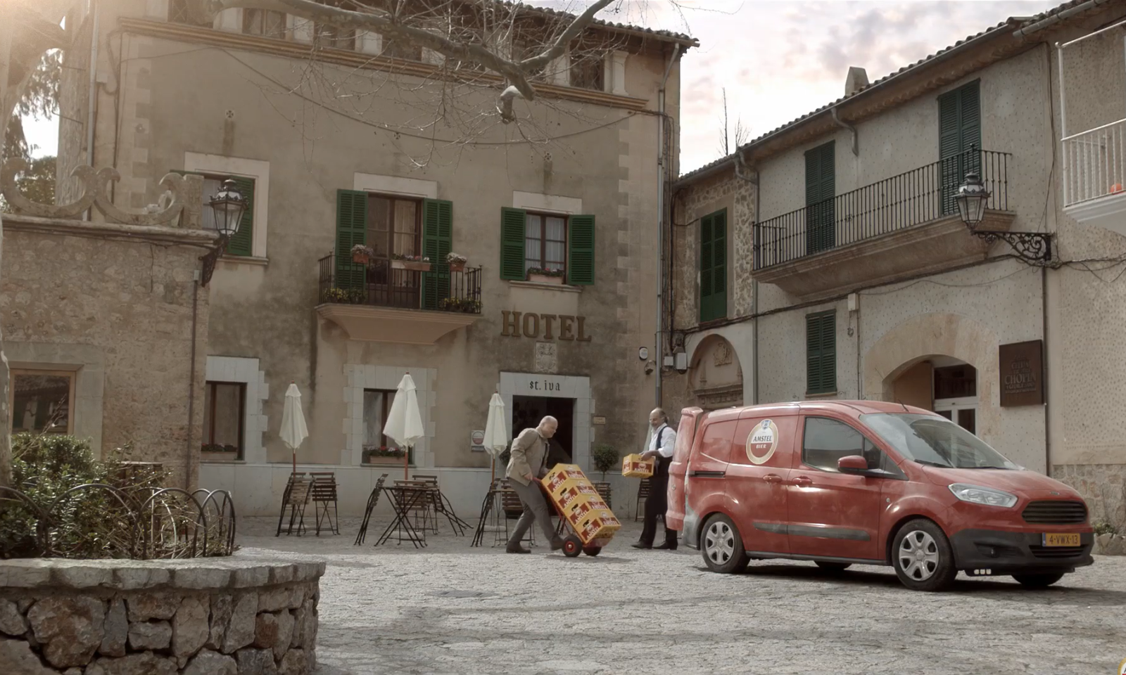 Twee mannen lossen dranken uit een busje voor een historisch hotel in een zonnig dorpsplein.