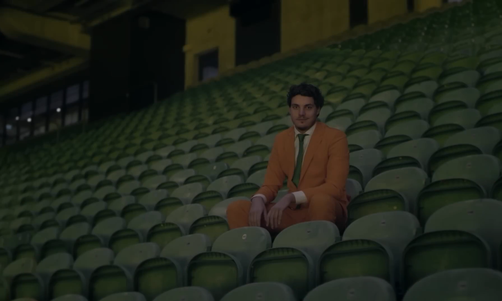 Man in oranje pak zittend op lege groene stadionstoelen