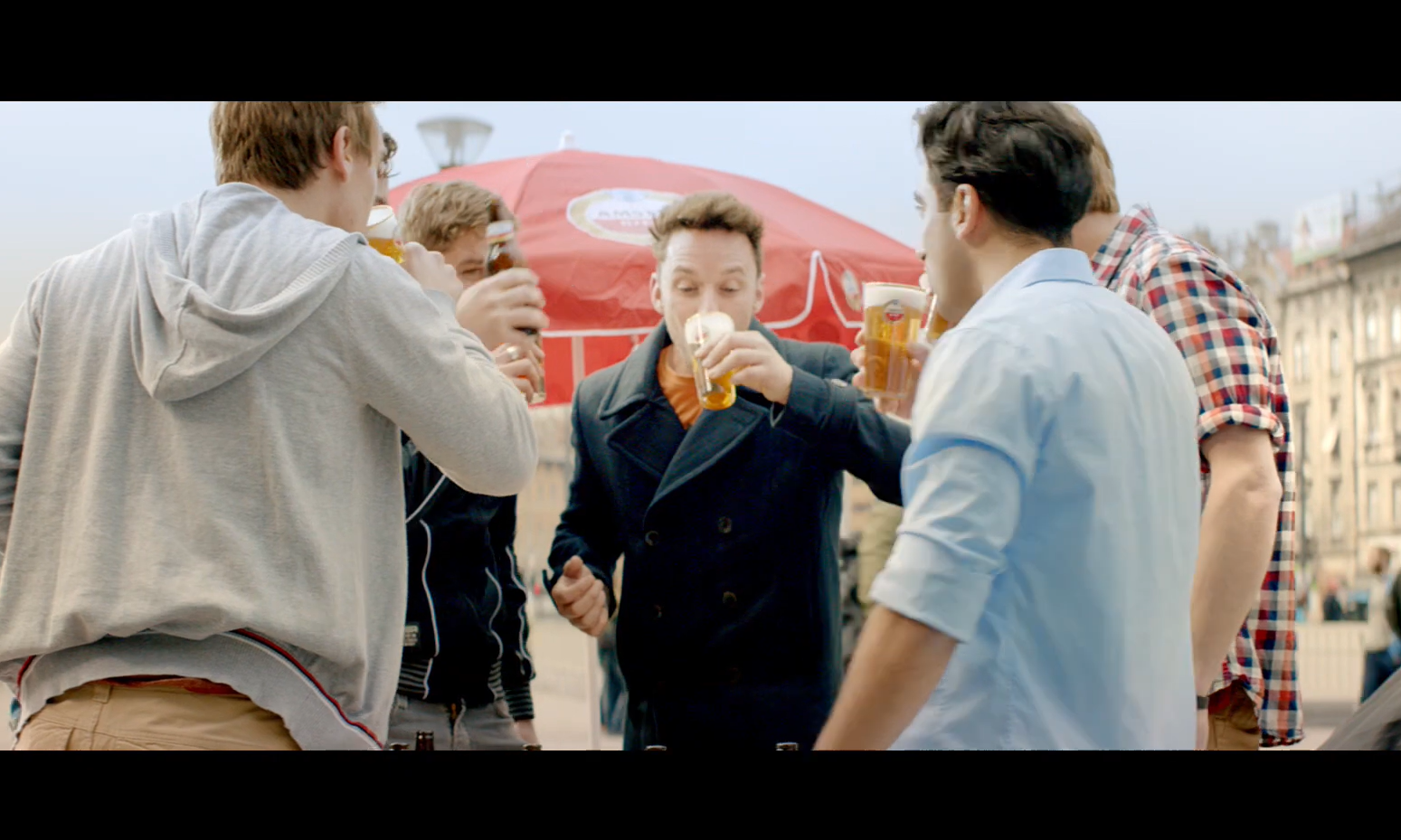 Vijf mannen drinken bier onder een rood parasol op een stadsplein.