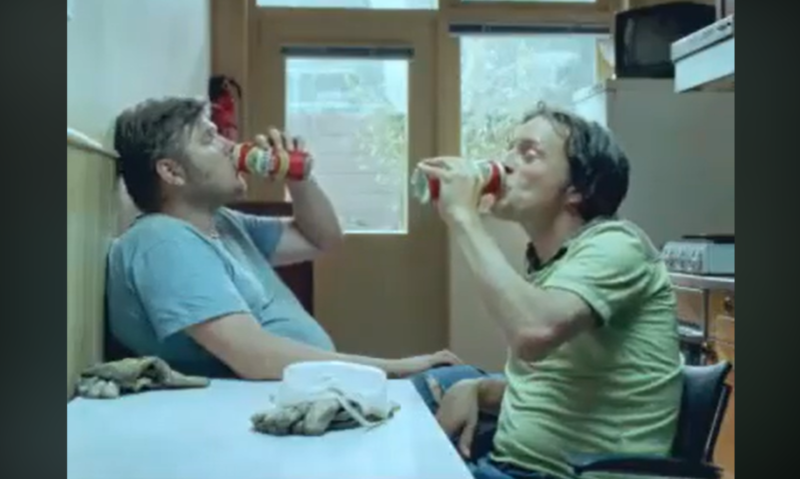 Twee mannen drinken bier in een keuken, zittend tegenover elkaar aan een tafel.