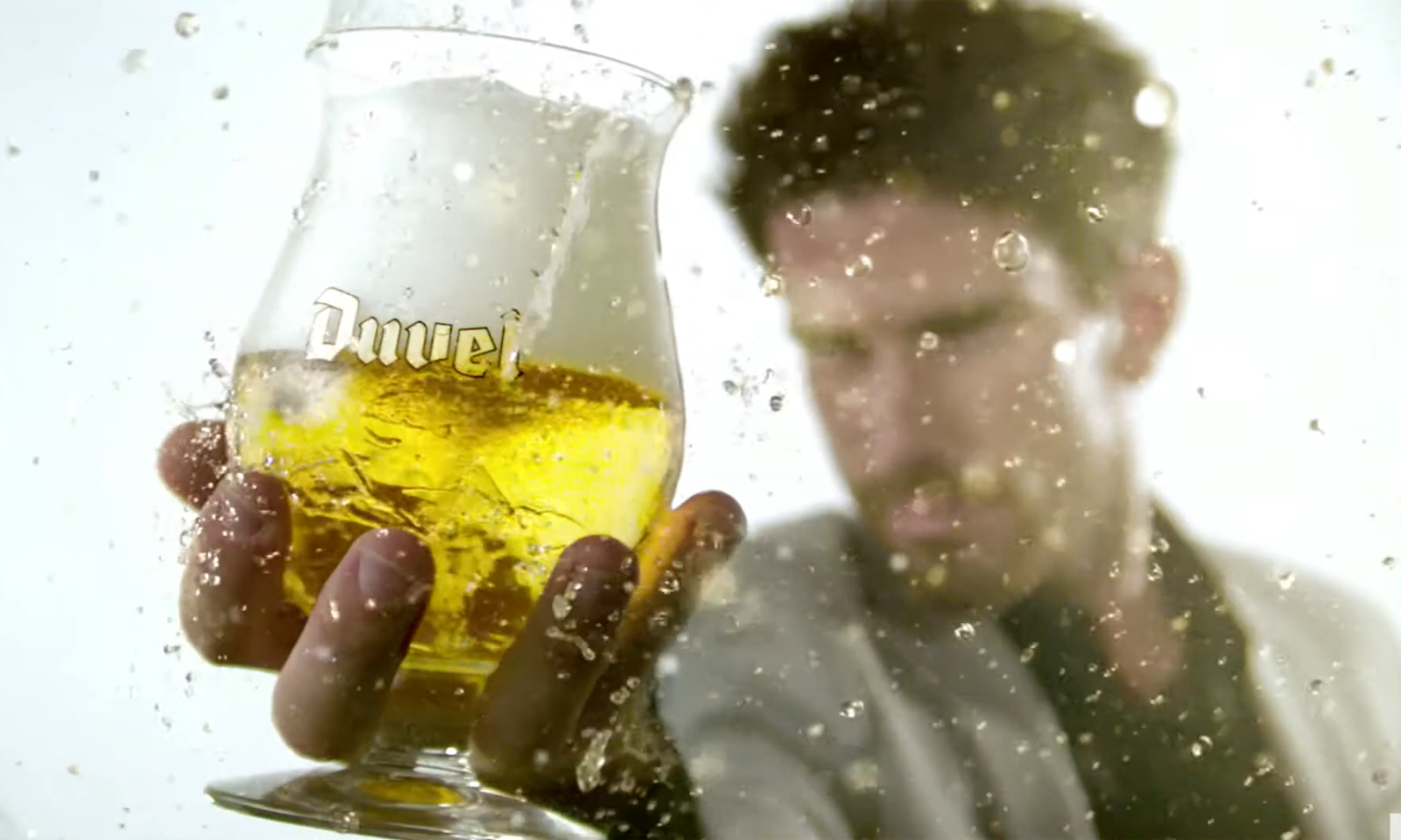 Hand houdt een Duvel glas met goudgele vloeistof en spetters in de lucht.