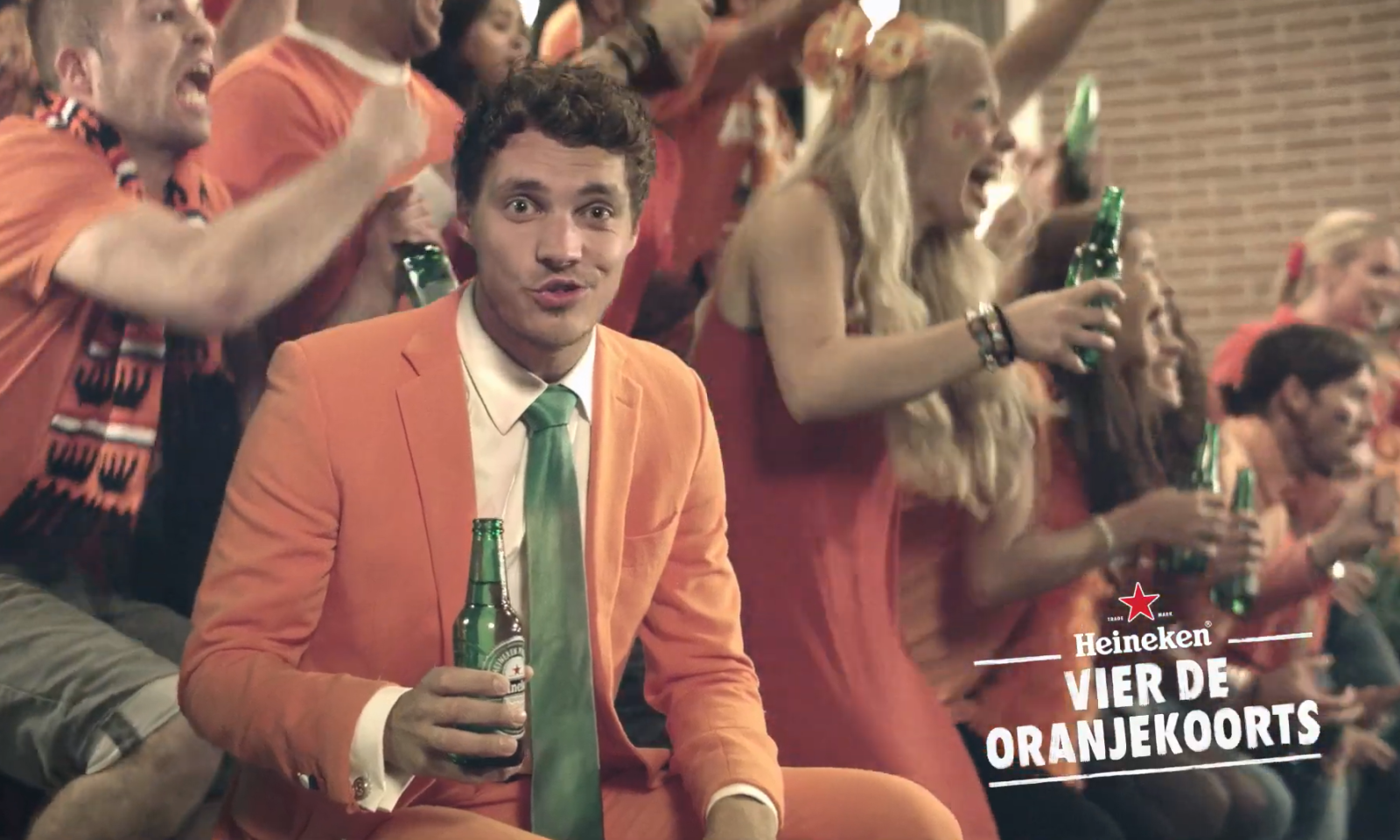 Man in oranje pak met groen-gestreepte das houdt een biertje vast te midden van juichende mensen.