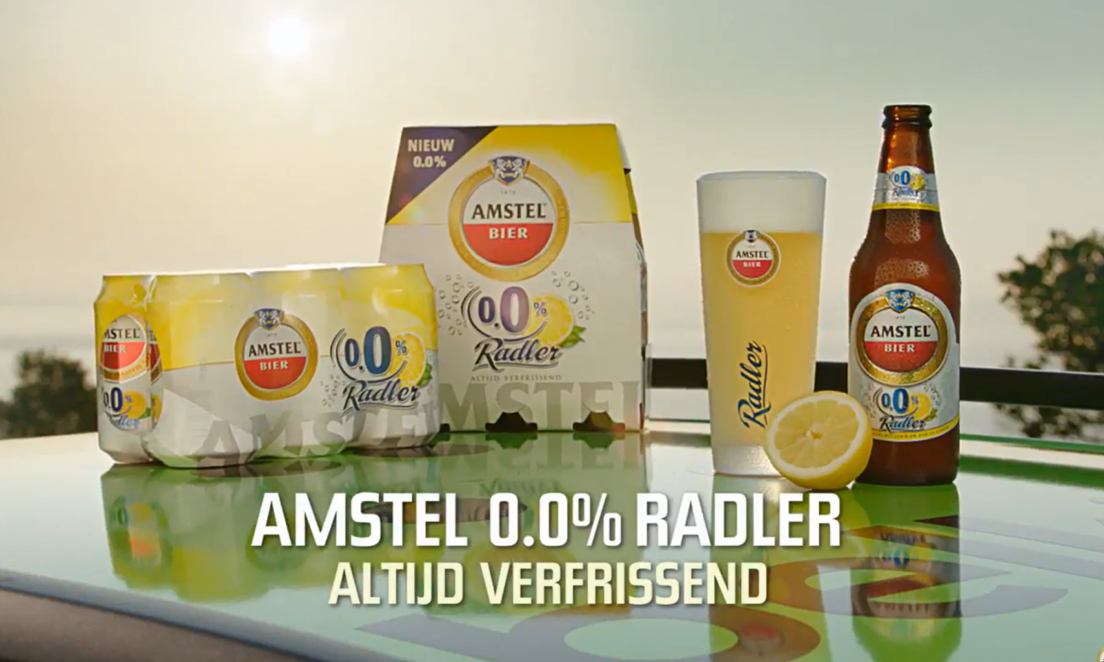 Pak Amstel 0.0% Radler, fles, glas bier en halve citroen op een tafel in de zon.