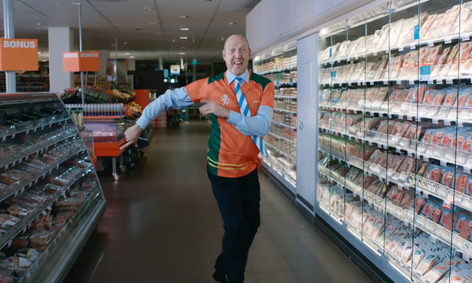 Man in Oranje Heineken sambashirt danst in supermarkt gangpad naast koelschappen.