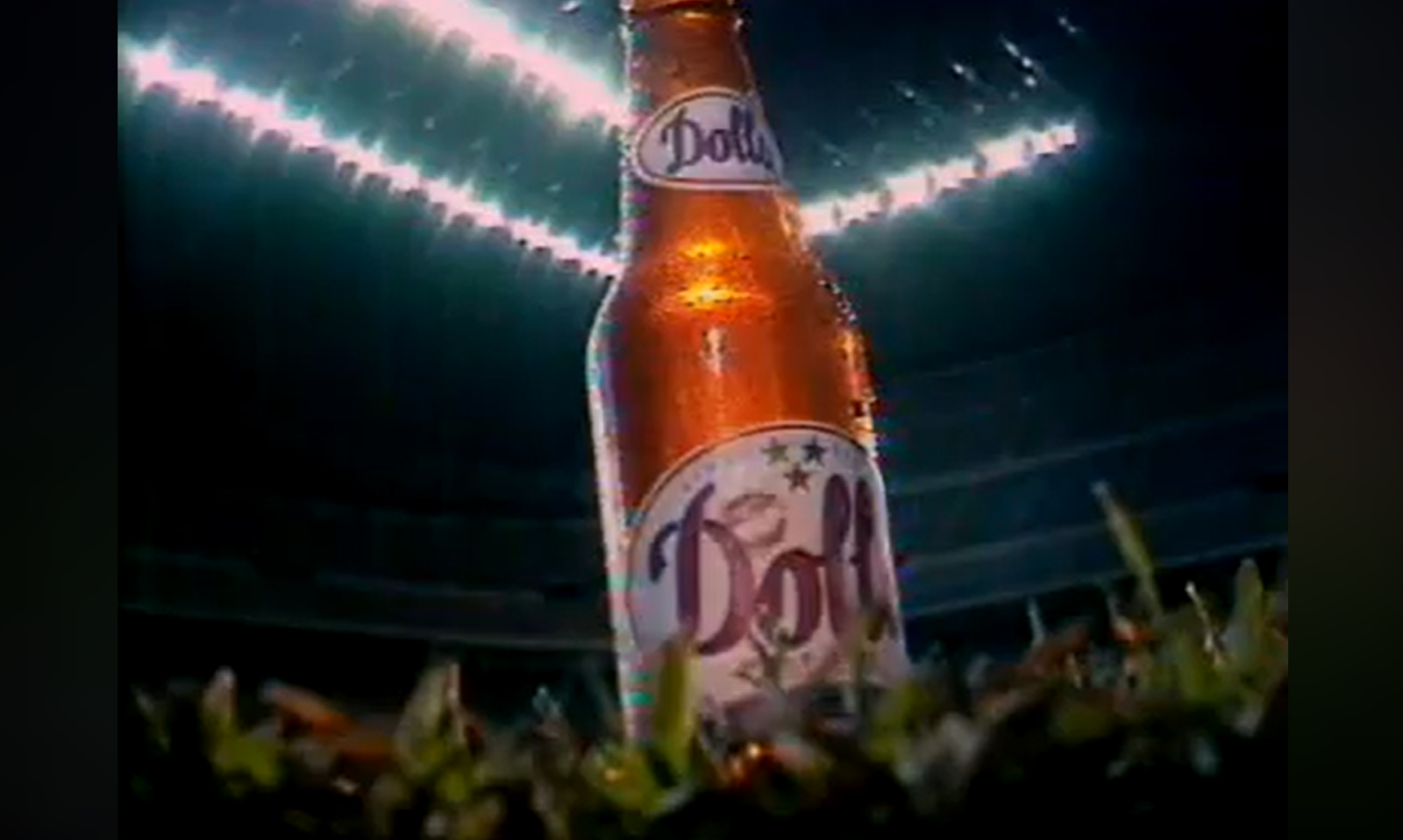 Een flesje Dolt-bier op een voetbalveld met stadionverlichting op de achtergrond.