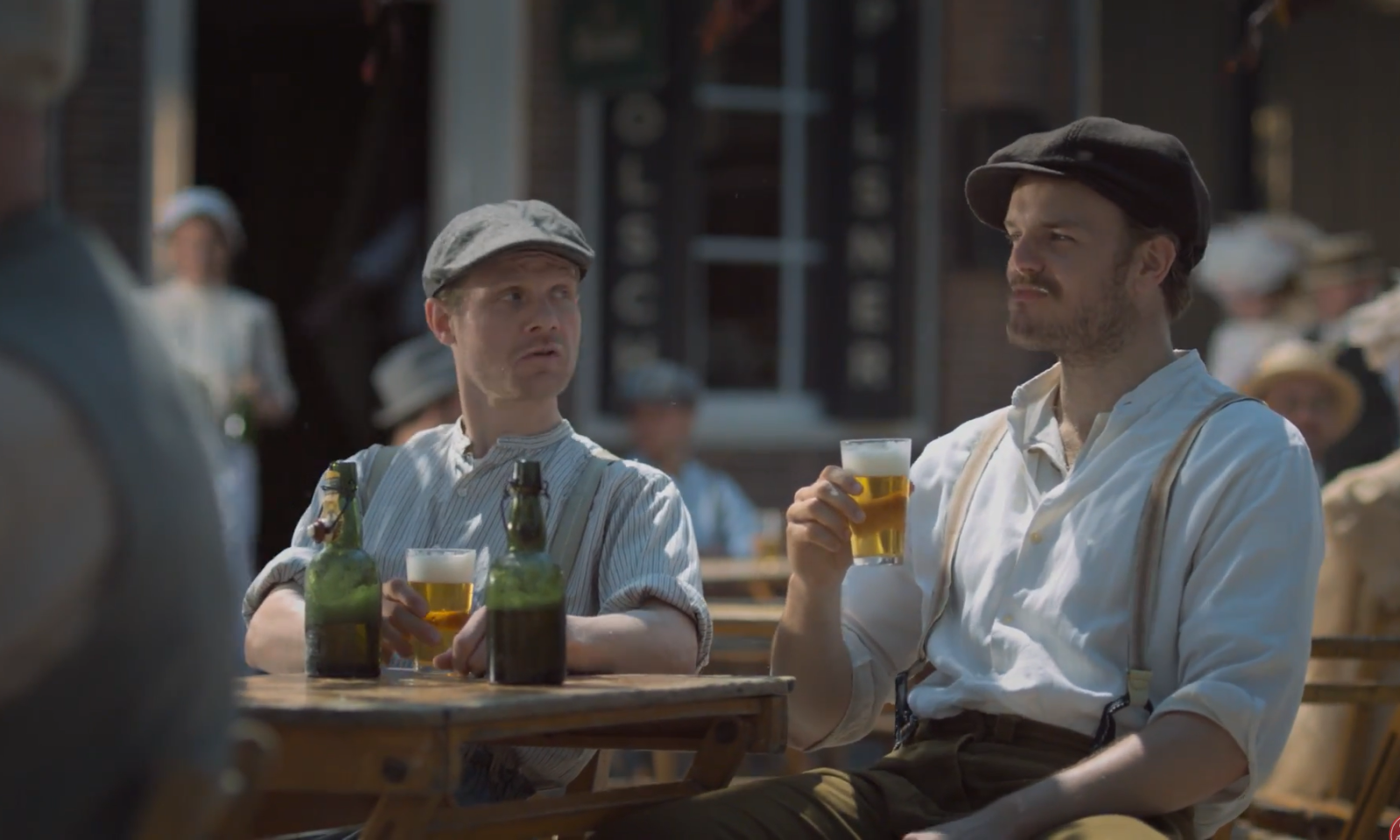Twee mannen in klassieke kleding zitten buiten met bier op een houten tafel.