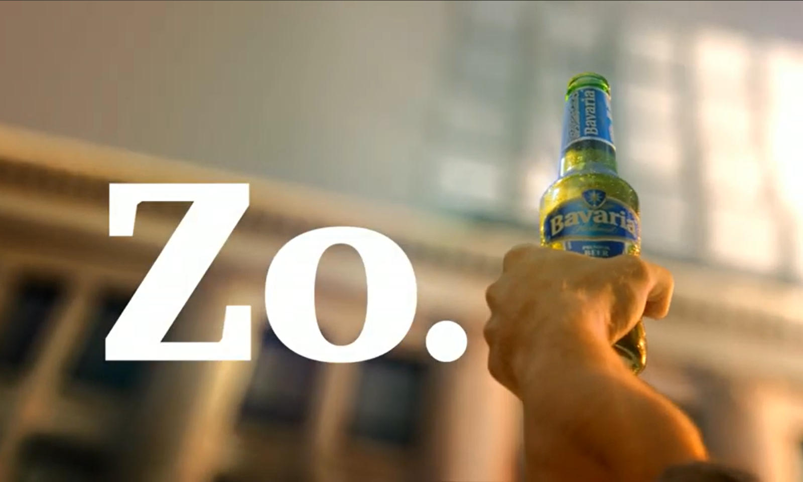 Hand houdt een flesje Bavaria bier omhoog tegen een wazige architecturale achtergrond.