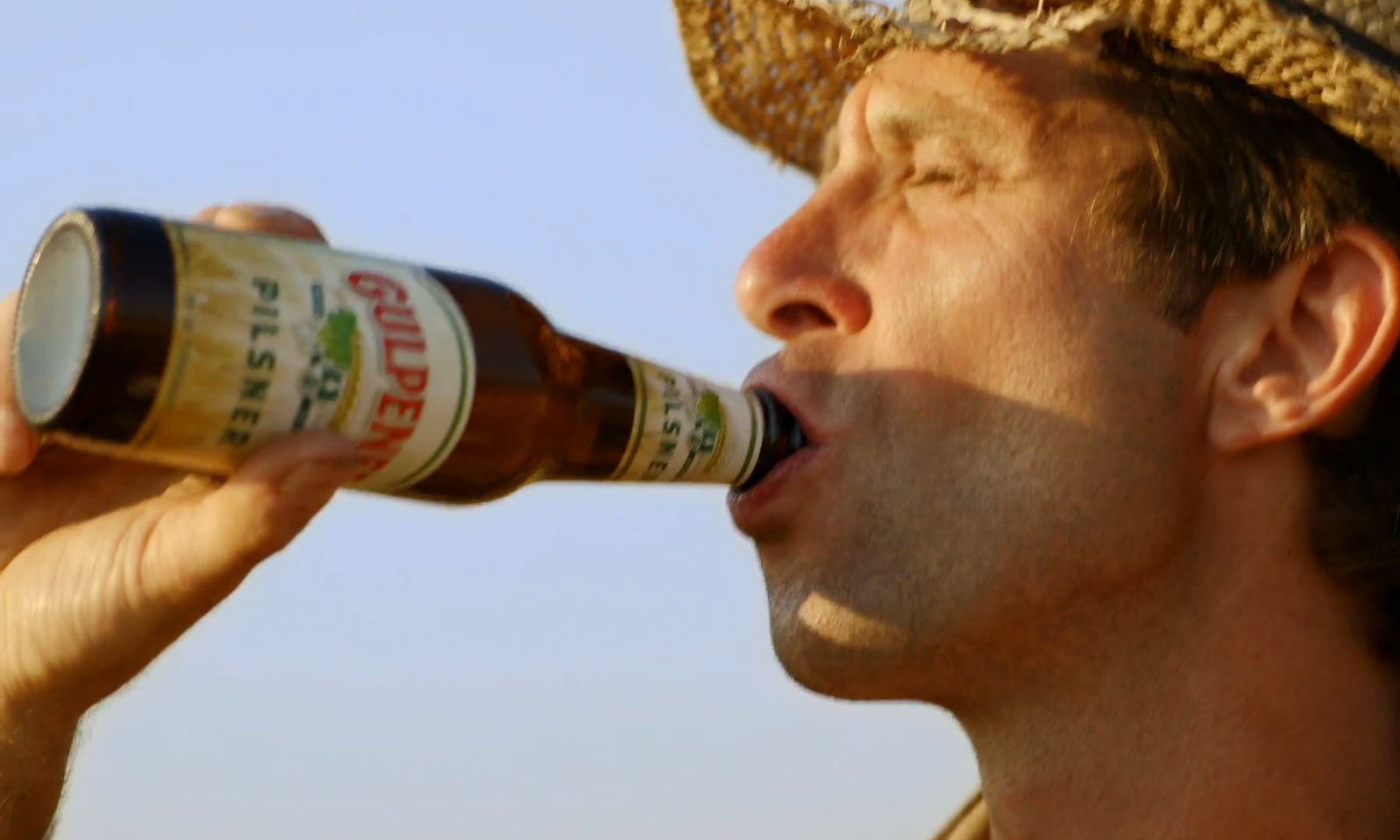 Man met strohoed drinkt een flesje pilsner onder een helderblauwe hemel.