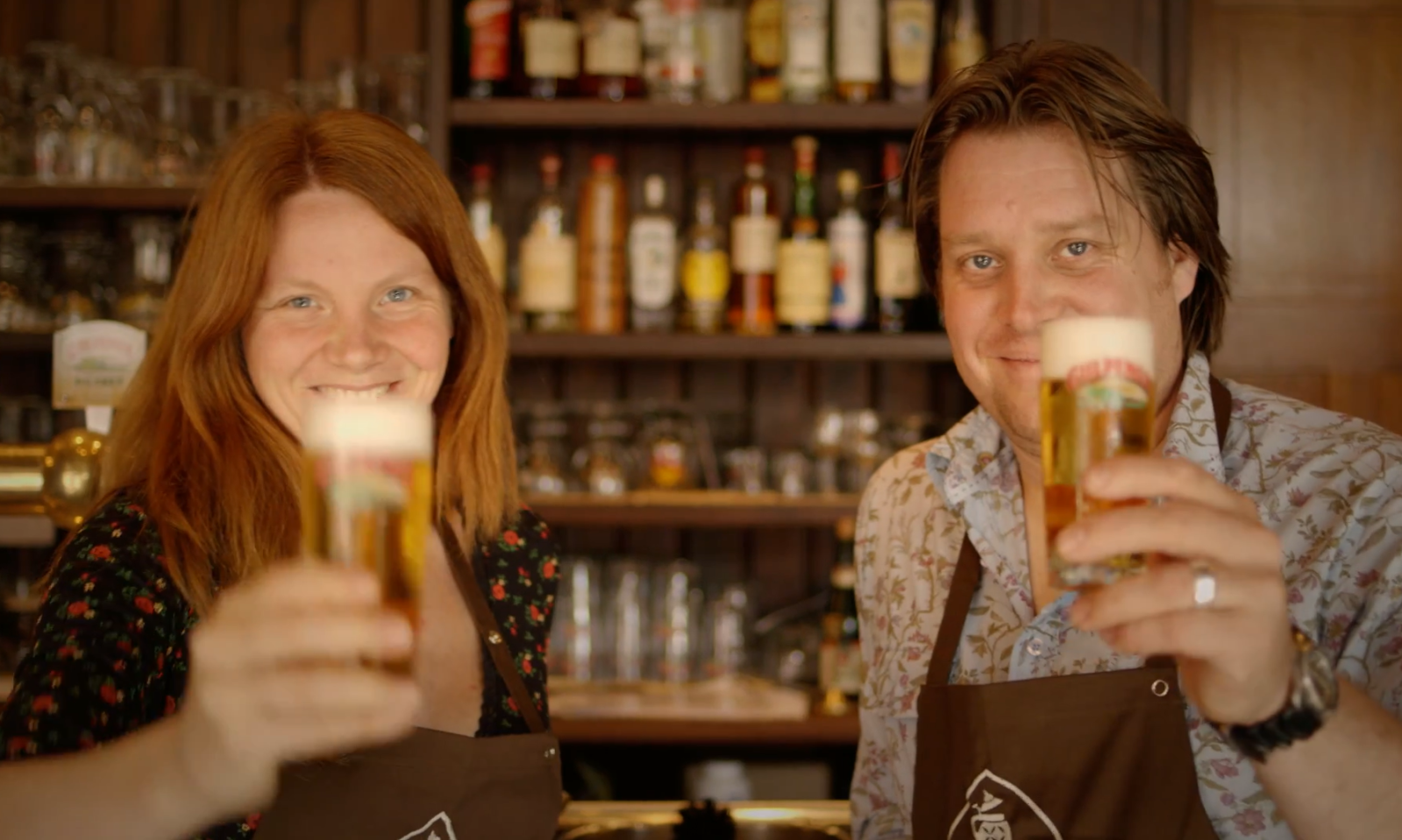 Twee mensen in een bar proosten met glazen bier voor een rek vol flessen.