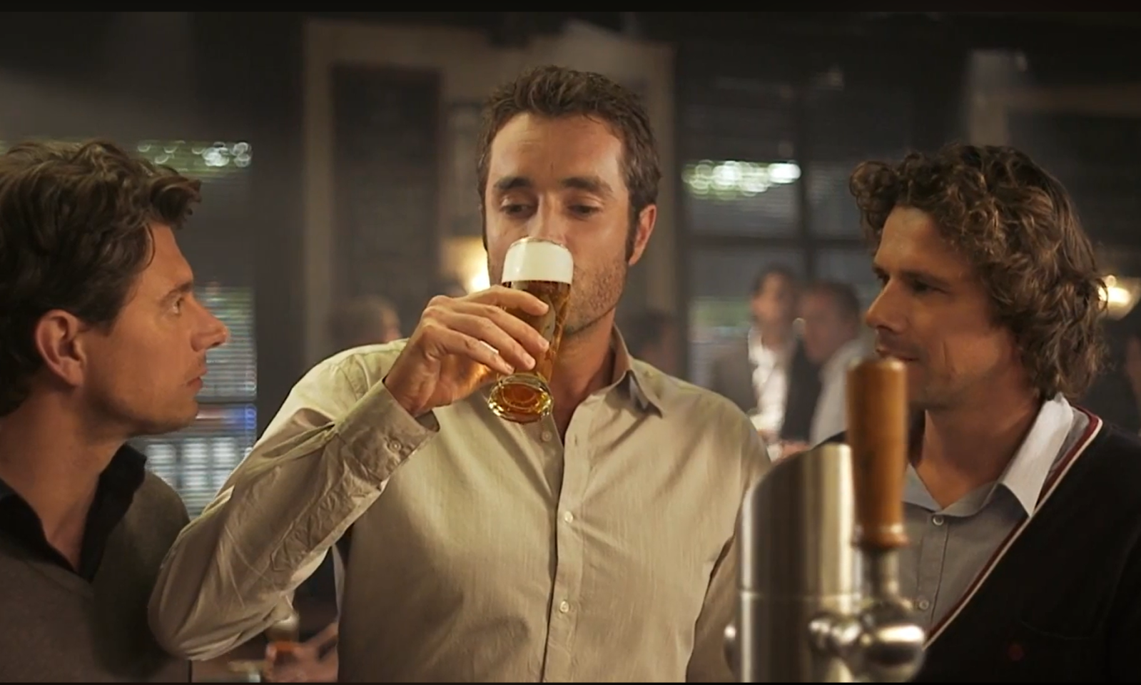 Drie mannen aan de bar, één drinkt een glas bier met schuimkraag.