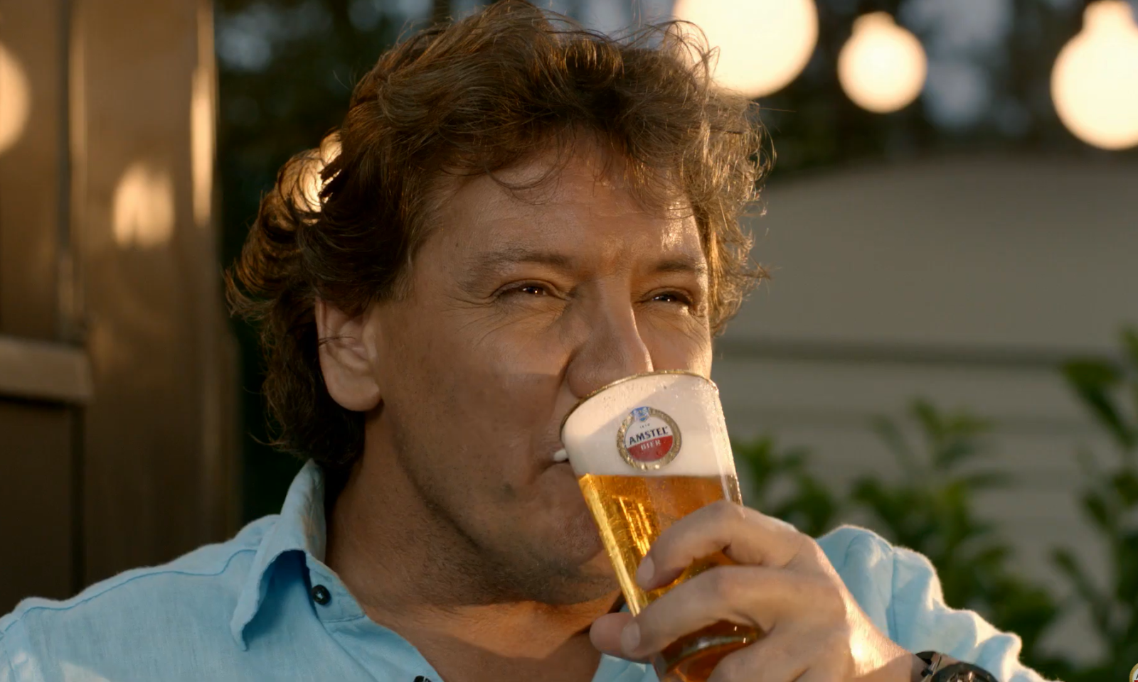 Man in een blauwe blouse drinkt een glas Amstel bier in een tuin setting.