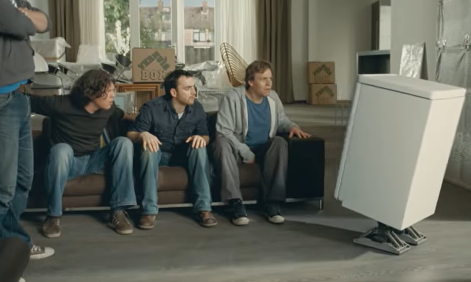 Een groep mannen op een bank kijkt verbaasd naar een koelkast die op robotschoenen staat.