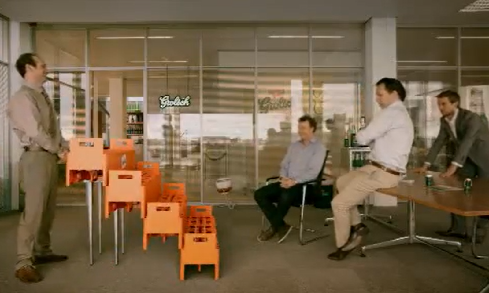 Vier mannen in een kantoor kijken naar een oranje bouwsel van bierkratten.