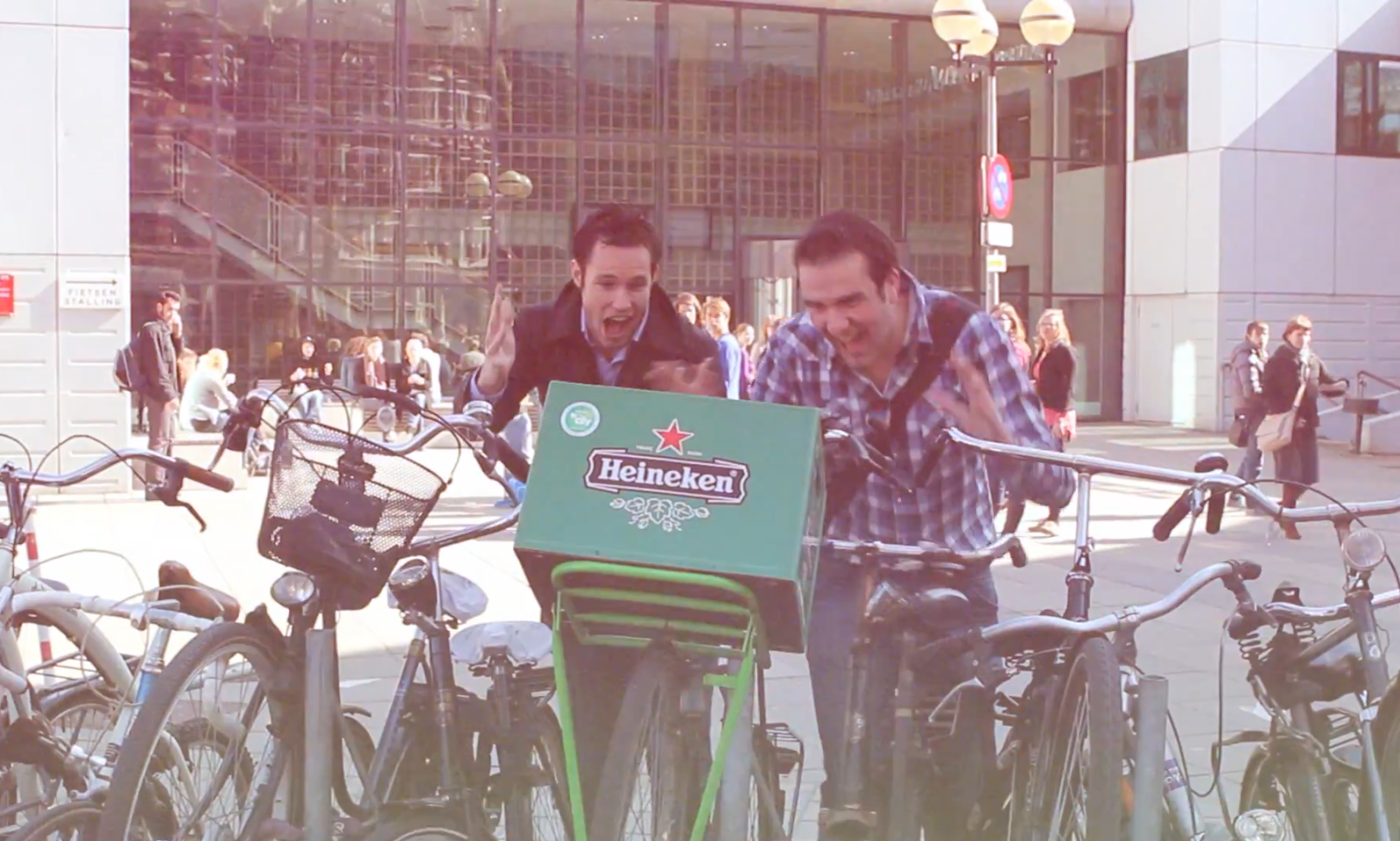 Twee mannen lachen en wijzen naar een fietskrat met Heineken logo buiten.