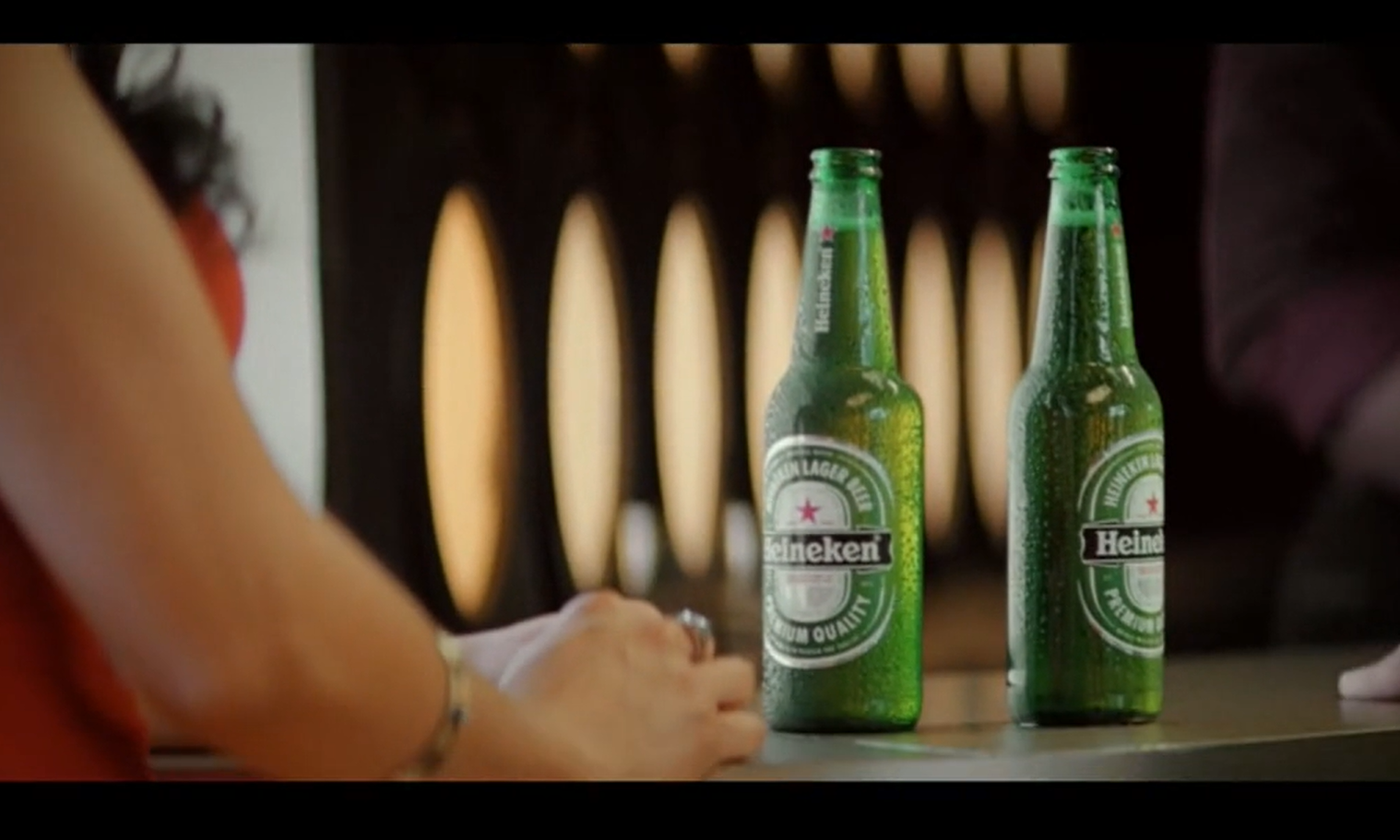 Twee Heineken bierflessen op een bar naast een persoon in een rode jurk.