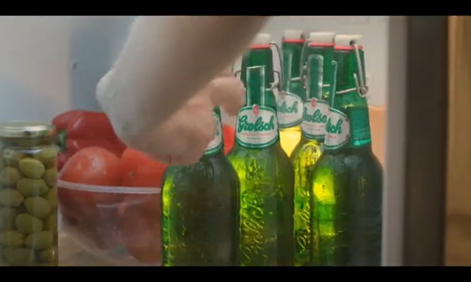 Groene bierflesjes in een koelkast naast een schaal met tomaten en een pot olijven.