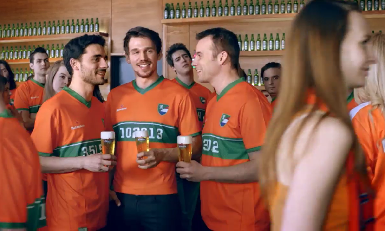 Mensen in oranje shirts praten en drinken bier in een bar met Heineken-flesjes op de achtergrond.