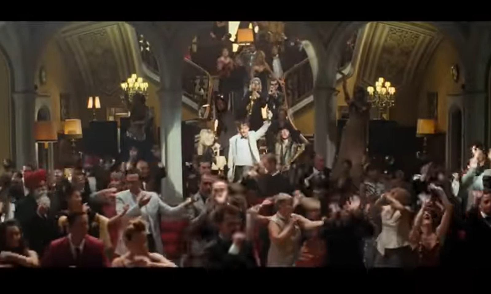 Mensen dansen in een luxueus ingerichte hal met een trap op de achtergrond.