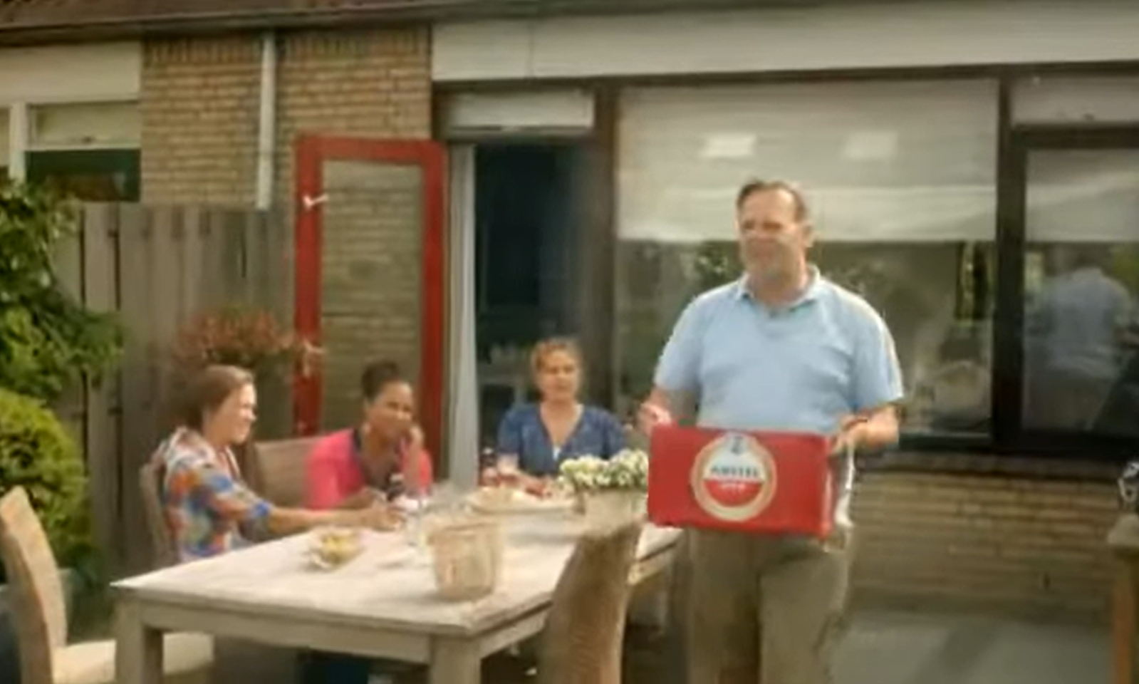 Man met krat bier bij tuinfeest met drie zittende mensen aan tafel.