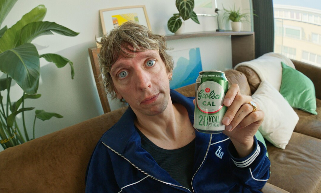 Faberyayo op een bank houdt een blik Grolsch CAL omhoog in een lichte woonkamer met planten.