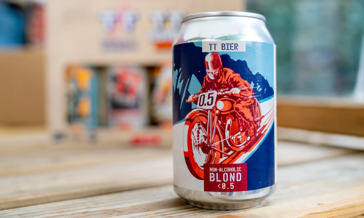 Blikje TT Bier Non-Alcoholic Blond 0.5% op houten tafel met wazige achtergrond van kartonnen doos en andere blikjes.