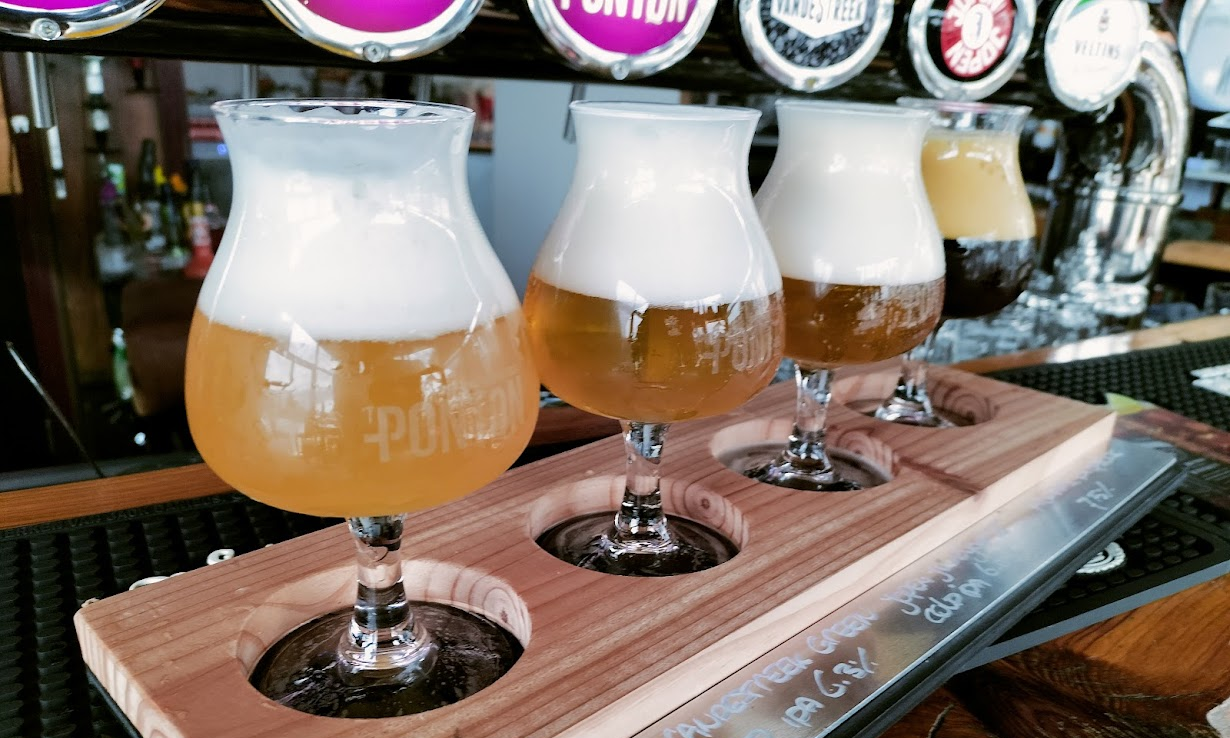 Vier bierglazen op een houten plank in een bar, met verschillende bieren.