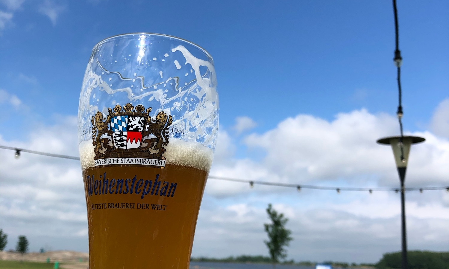Glas bier met schuim, bierlogo zichtbaar, tegen een blauwe lucht met enkele wolken.
